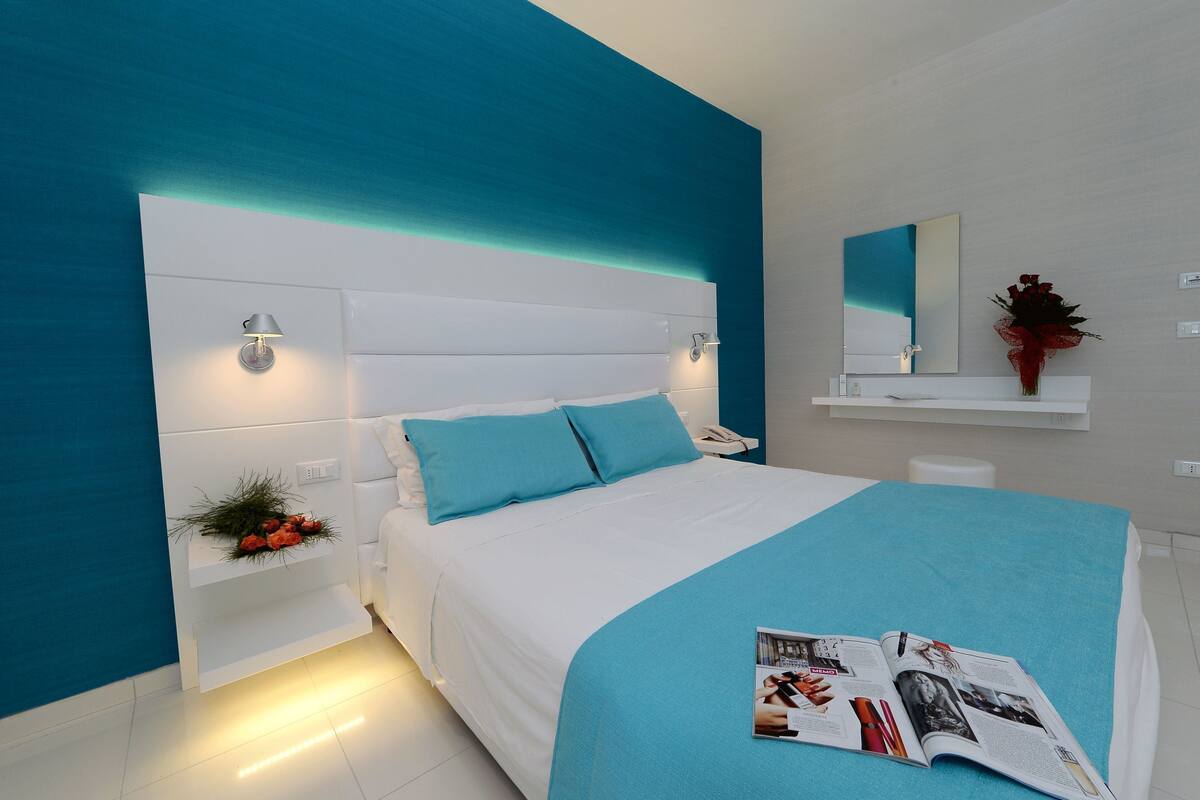 Une chambre moderne avec un lit blanc, des murs d’un bleu-vert vif, une table de chevet avec une lampe et un magazine posé sur le lit.