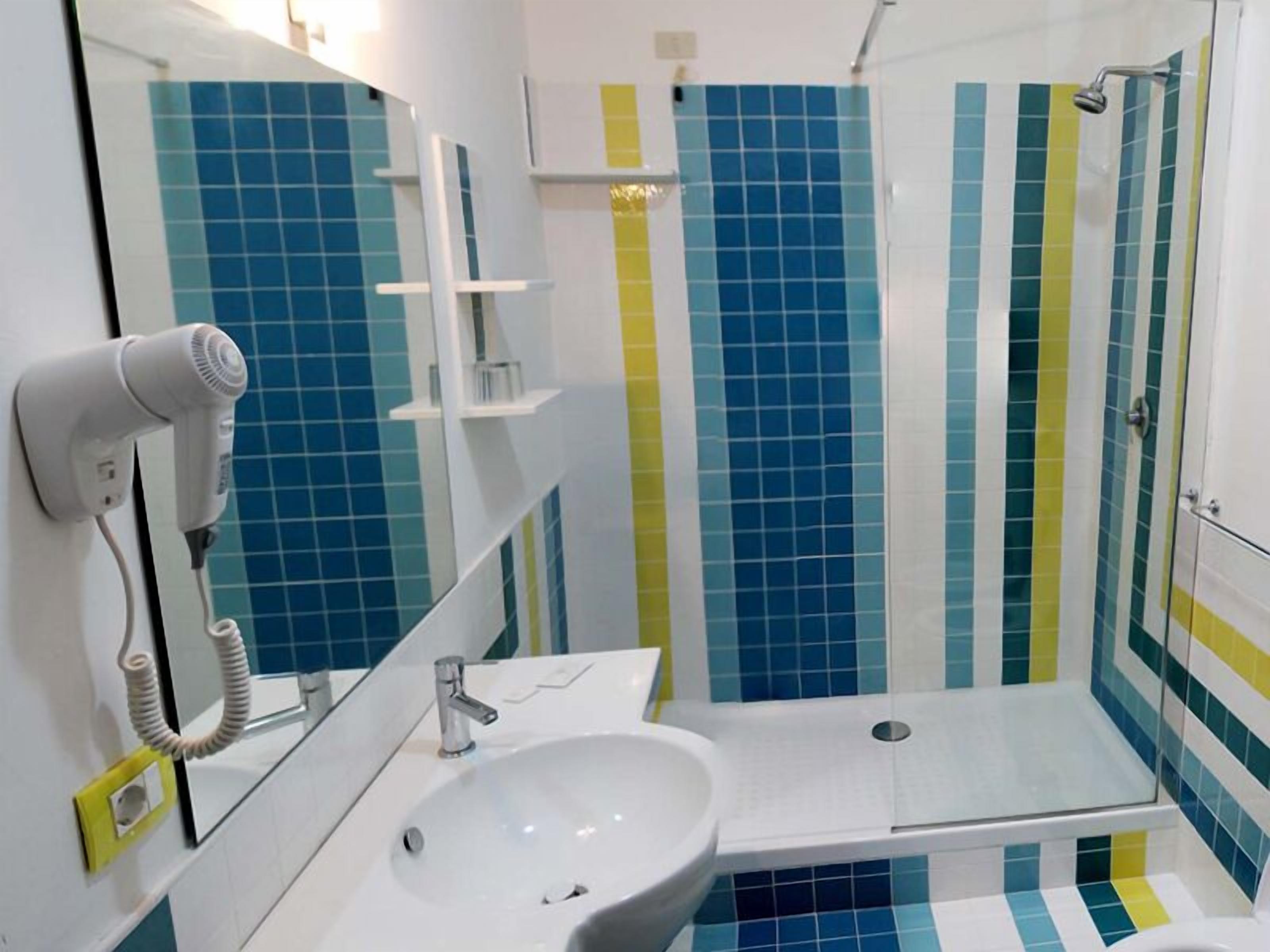 Une salle de bain avec un lavabo blanc, un miroir, un sèche-cheveux et une douche dont les carreaux sont bleus et jaunes.