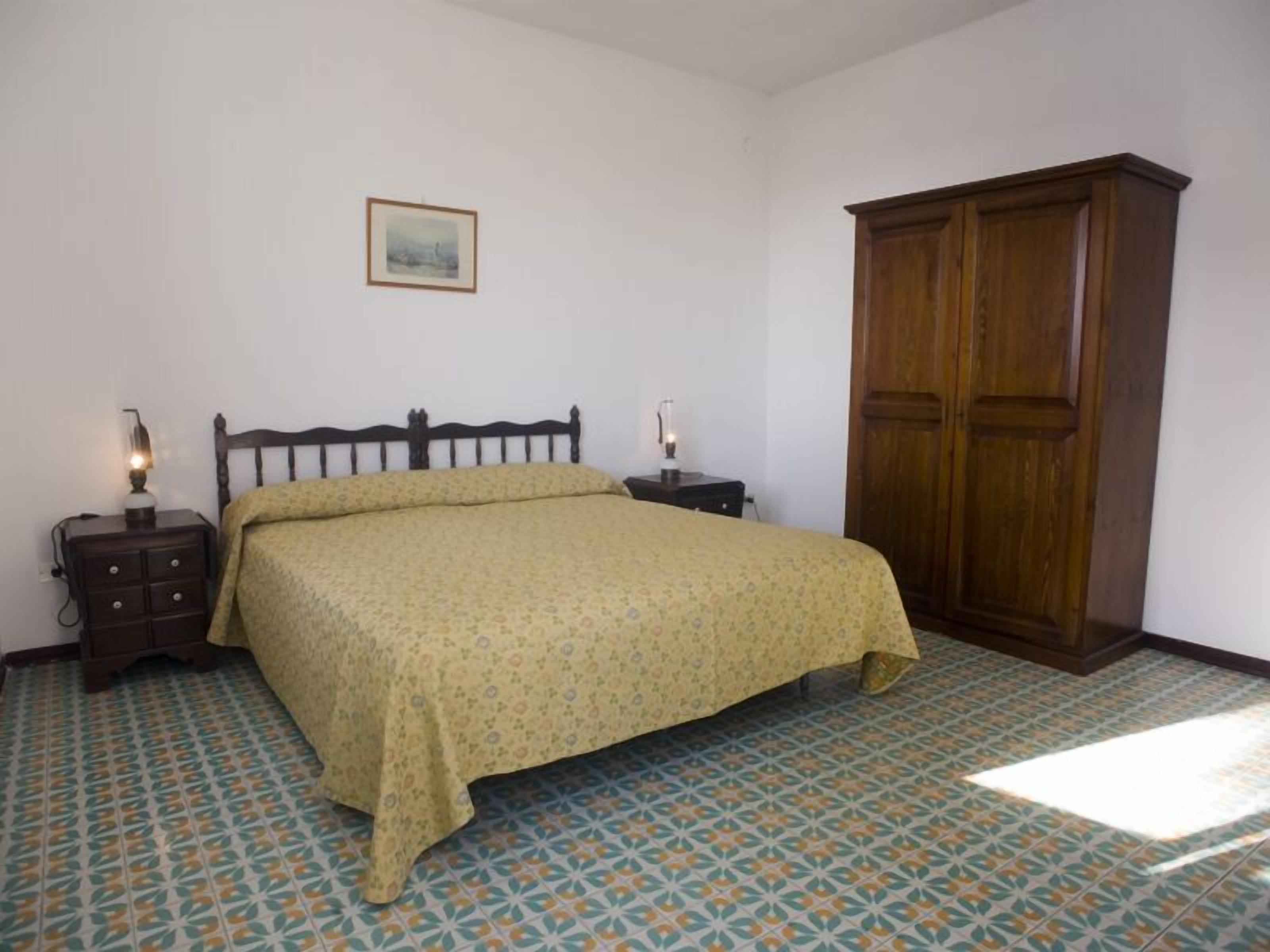 A bedroom with a bed, nightstands, a wardrobe, and patterned floor tiles.