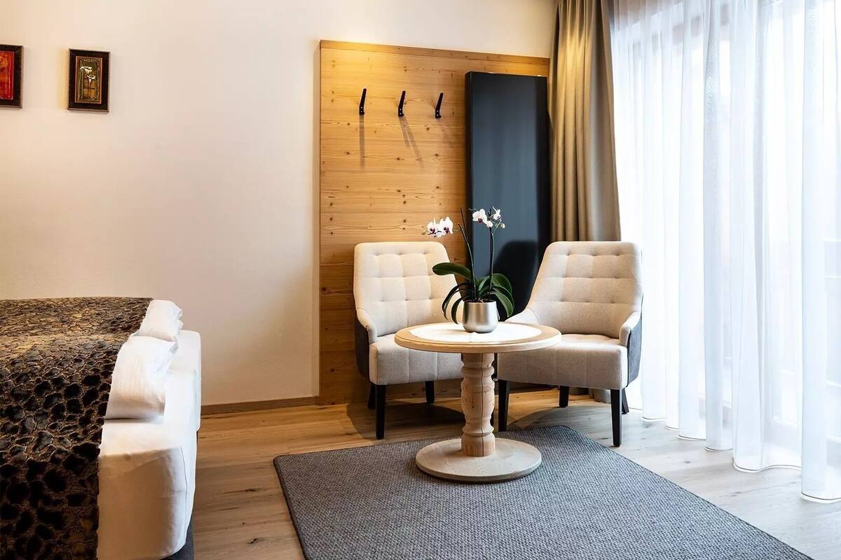 A modern hotel room with a wooden accent wall, two upholstered chairs, a small round table with a potted plant, and a bed with a patterned bedspread.