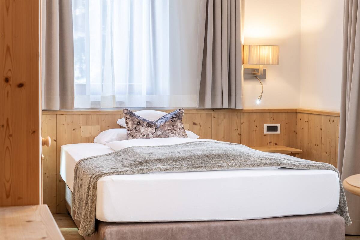 A hotel room with a wooden headboard, a bed with white linens, a patterned pillow, a bedside lamp, and a window with sheer curtains.
