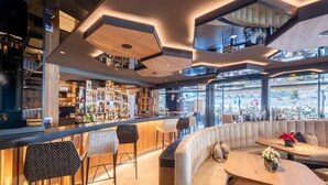 A modern bar with a high ceiling, bar stools, a seating area with a sofa and tables, and a view of the street through large windows. - Hotel Arkadia - Adults Only (Corvara in Badia)