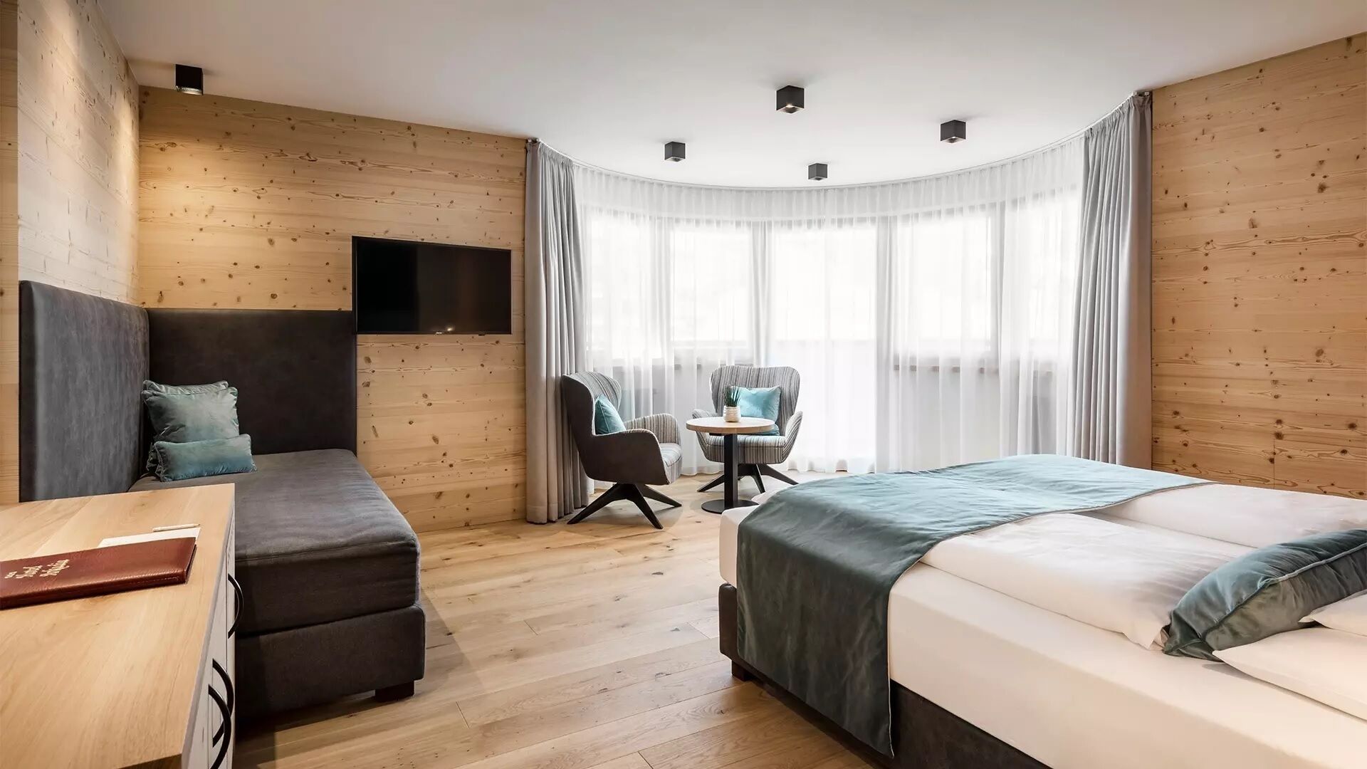 Una camera d'albergo moderna con parete in legno, televisione a schermo piatto, divano, letto, un piccolo tavolo e una sedia.