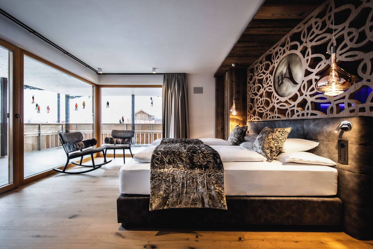 A modern bedroom with a large bed, a view of a ski slope, and a wooden ceiling.