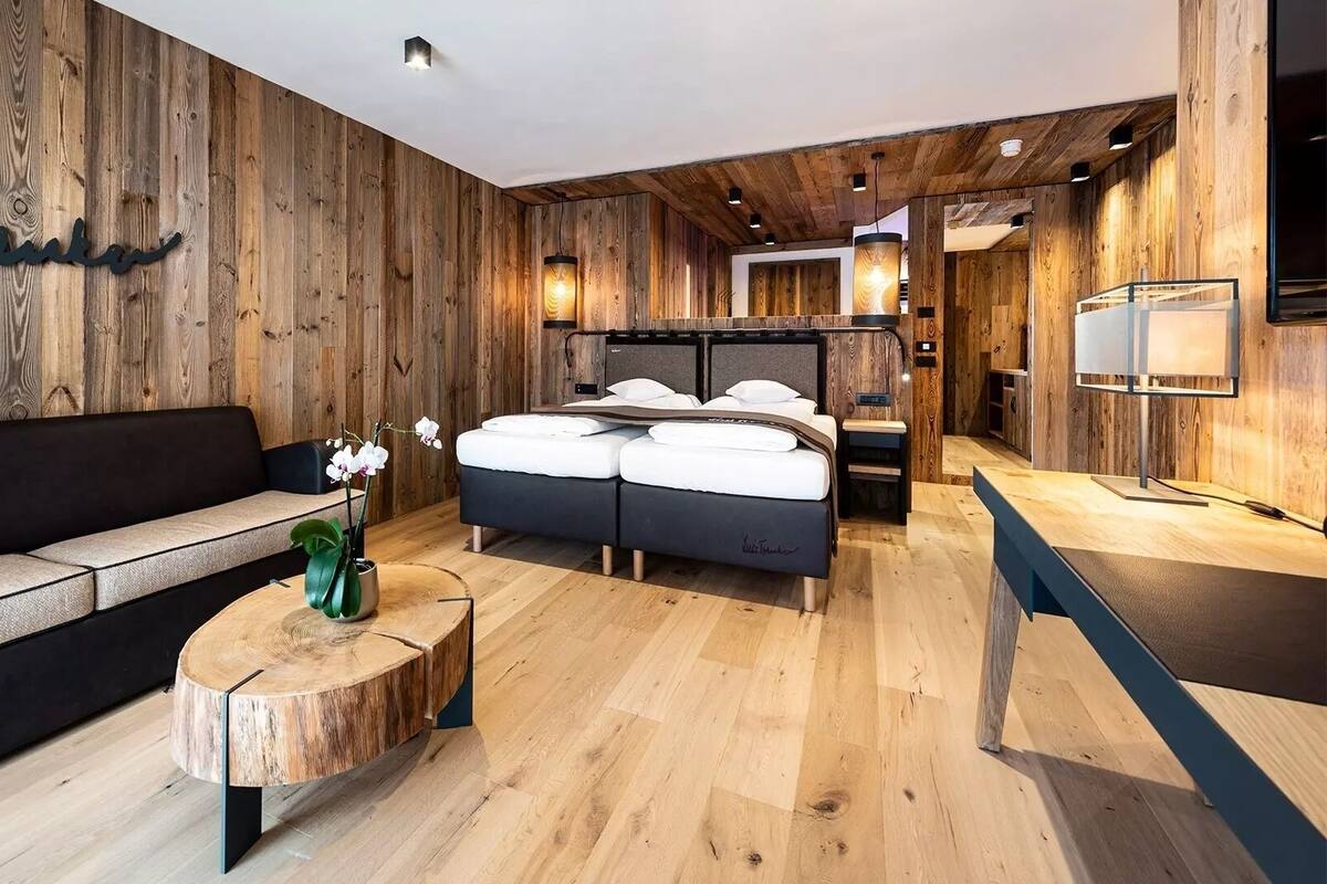 A modern hotel room with a wooden wall, a sofa, a bed, a round wooden coffee table, and a small table with a lamp.