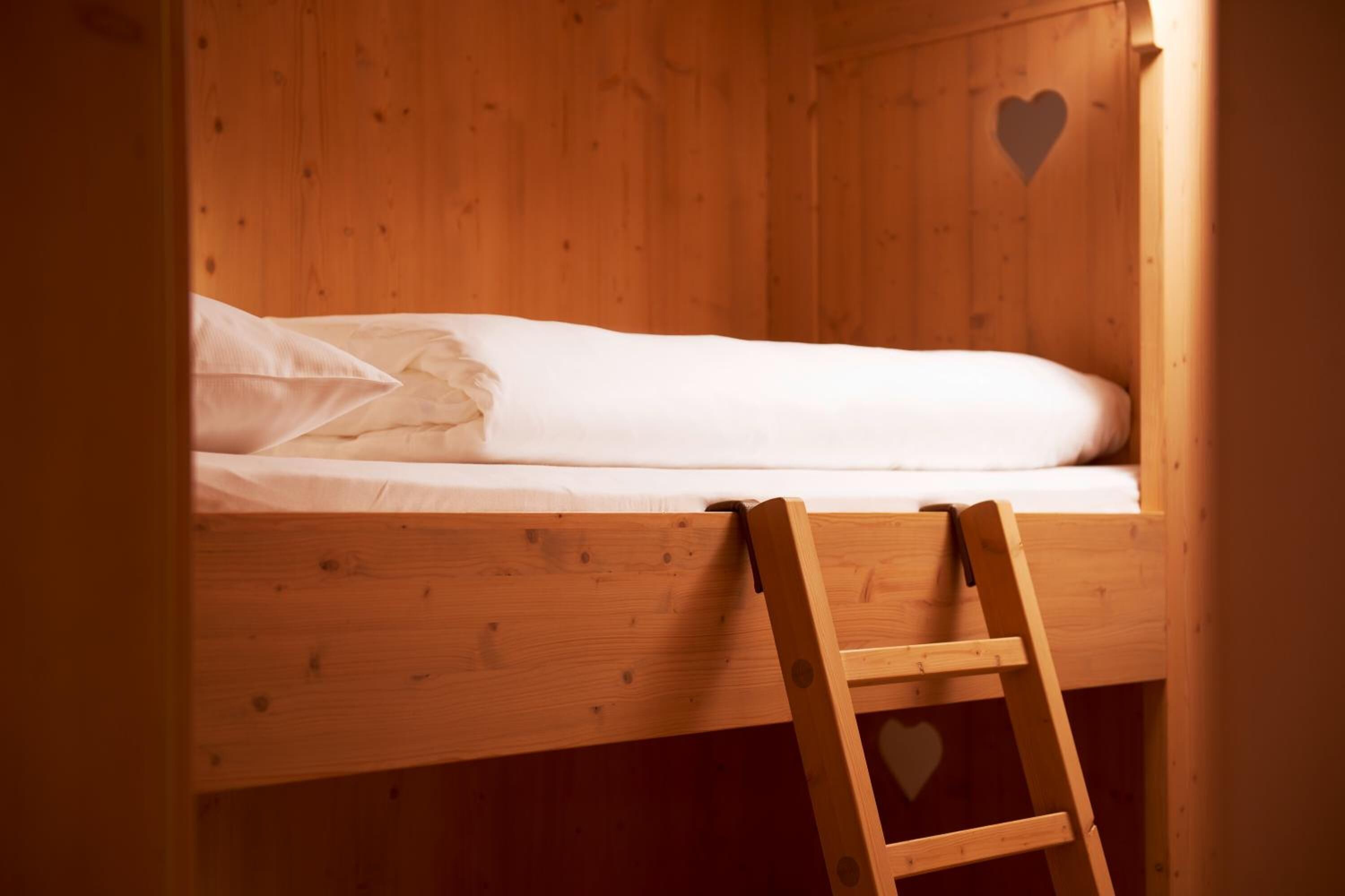 Ein Holzbett mit Etagen und einer Leiter, ein herzförmiges Loch in der Wand sowie ein weißes Bettlaken.