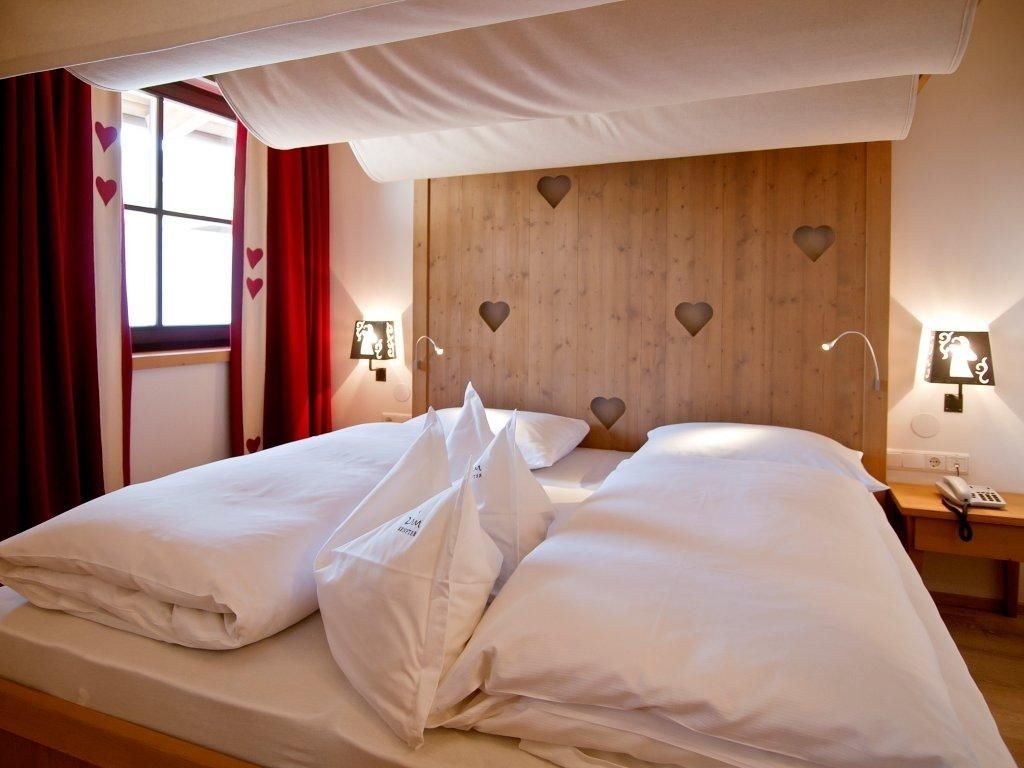 Un lit bien fait, avec du linge de lit blanc et des oreillers, une tête de lit en bois ornée de découpes en forme de cœur, des lampes de chevet et une fenêtre avec des rideaux rouges.