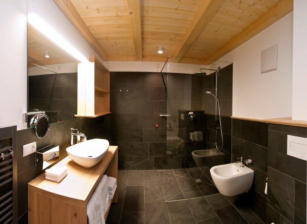 A modern bathroom with a wooden ceiling, a freestanding sink, a bidet, and dark tiled walls.