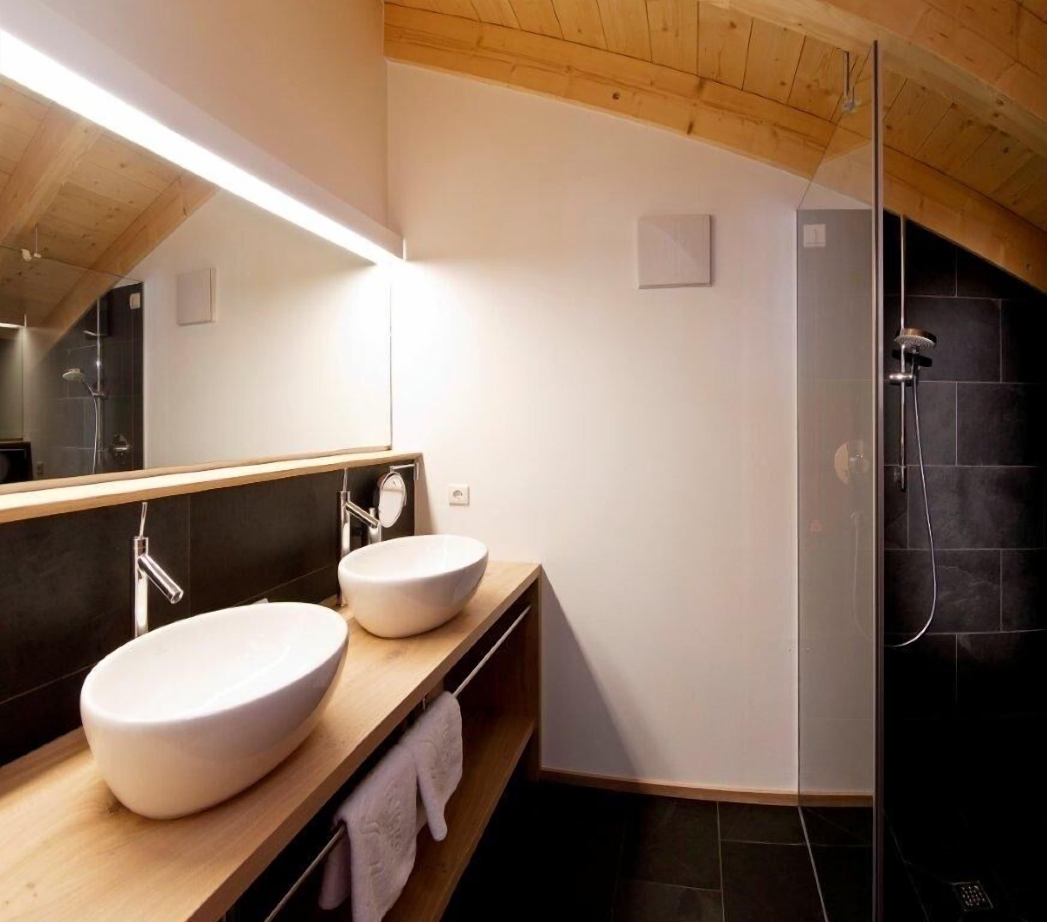 A modern bathroom with two large white sinks, a wooden vanity, a large mirror, and a glass-enclosed shower area.
