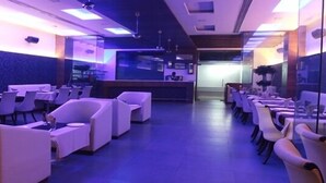 Un moderno vestíbulo de hotel con un ambiente iluminado en azul, que cuenta con una recepción, varias áreas de estar y una cocina visible al fondo.