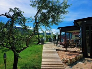 A wooden deck leading to a covered outdoor seating area with tables and chairs, surrounded by greenery and a clear sky. - Antico Fienile Agritur (Mezzocorona)