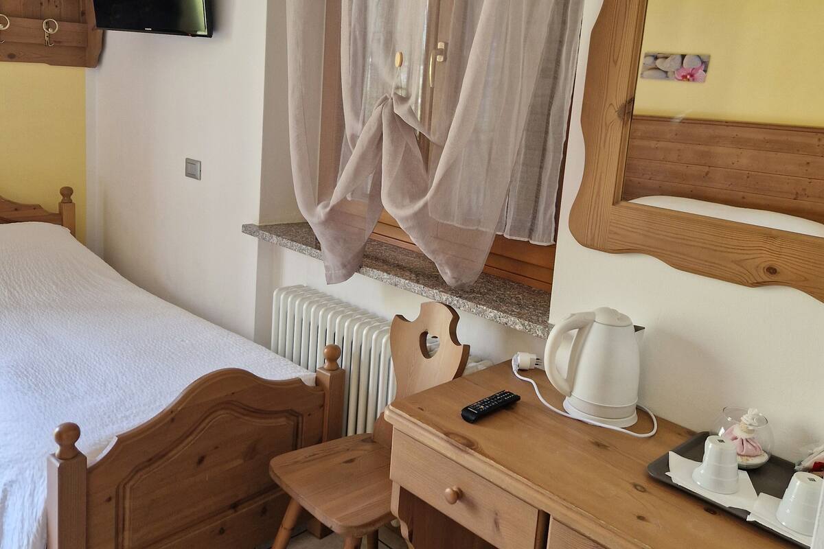 A bedroom with a bed, a wooden desk, a chair, a television, a window with curtains, a radiator, and a wooden cabinet.