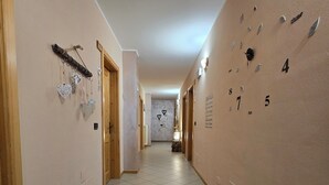 A hallway with a clock on the right wall, a door on the left, and a small table with a vase in the distance.
