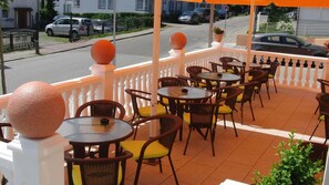 A covered outdoor seating area with wicker chairs and tables, a white railing, and a view of the street. - Hotel Selliner Hof (Sellin)
