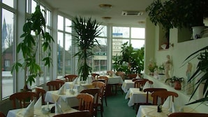 Un restaurante con mesas dispuestas para comer, ventanas amplias y diversas plantas en macetas.