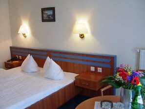 A hotel room with a bed, two bedside lamps, a nightstand, a small table with a vase of flowers, and a wall-mounted picture. - Hotel Selliner Hof (Sellin)