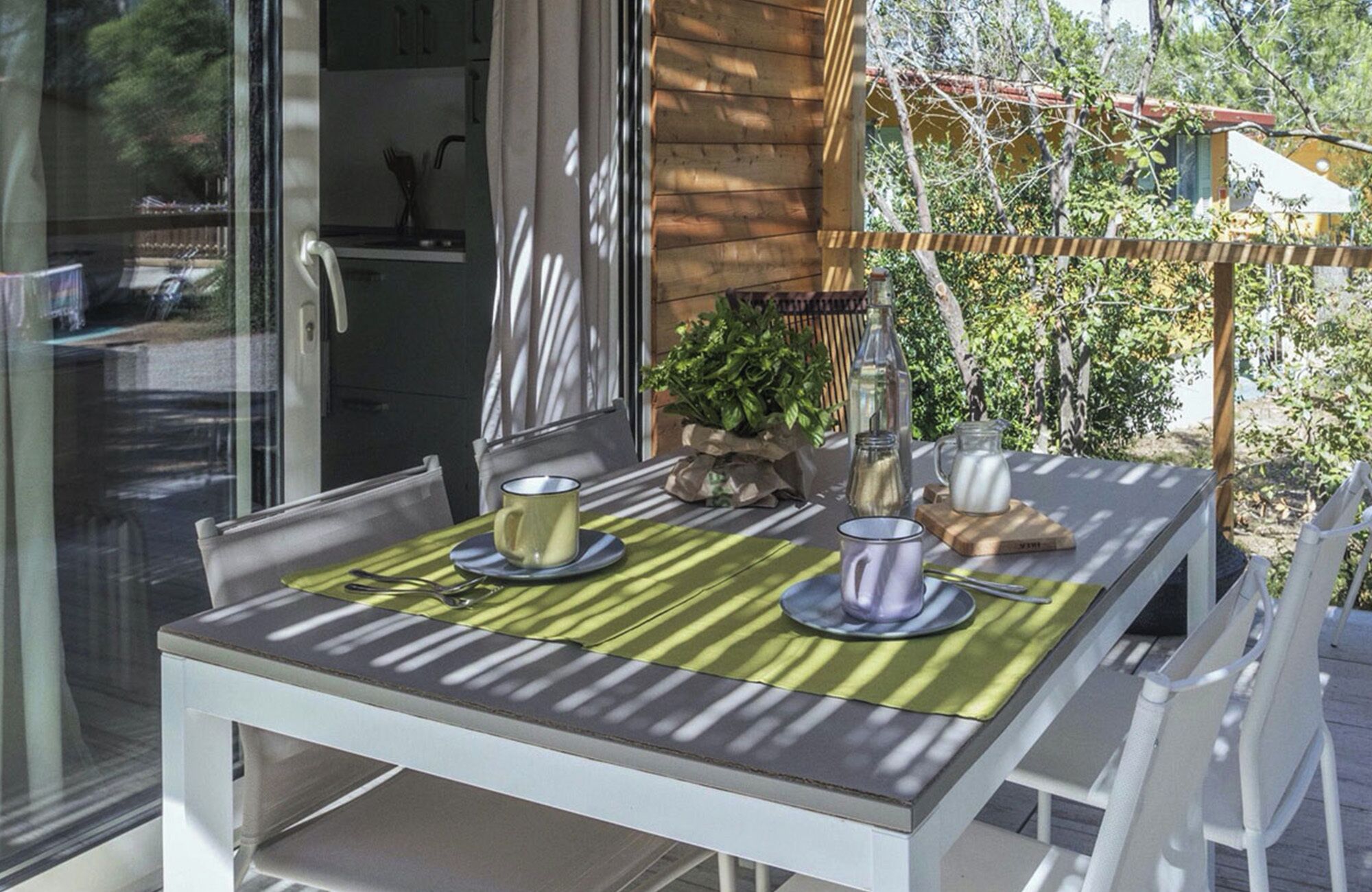 Une table à manger avec une nappe à rayures vertes et blanches, deux tasses et un pichet, le tout posé sur une table en bois à l’extérieur.