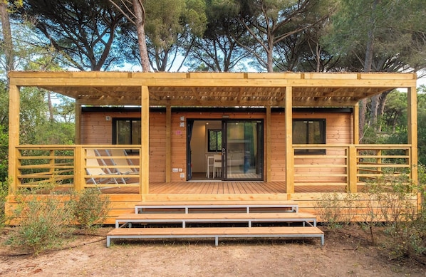 A wooden cabin with a deck, a glass door entrance, and a small table and chair inside.