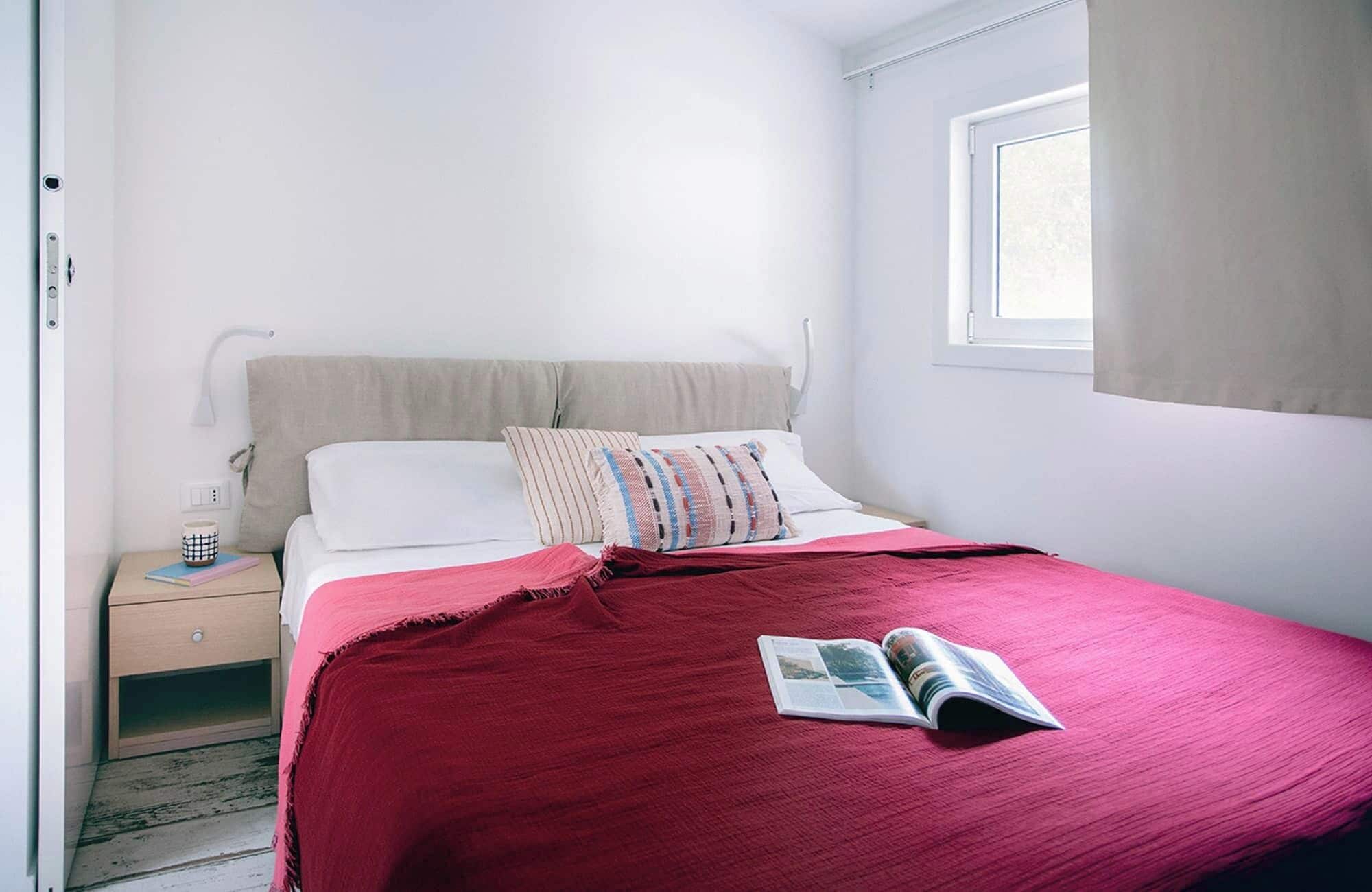 A bedroom with a bed, a nightstand, a window, and a magazine on the bed.