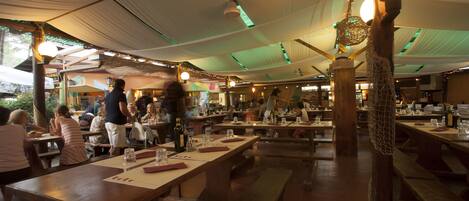 Un comedor interior con mesas y bancos de madera, techo alto con luces colgantes y suelo de ladrillo.