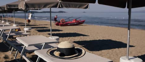 Beach chairs under umbrellas, a red boat on the water, and a person in the distance.