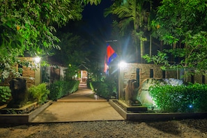 A pathway leading to a building with a flag, surrounded by greenery and stone statues. - Bunwin Residence (Siem Reap)