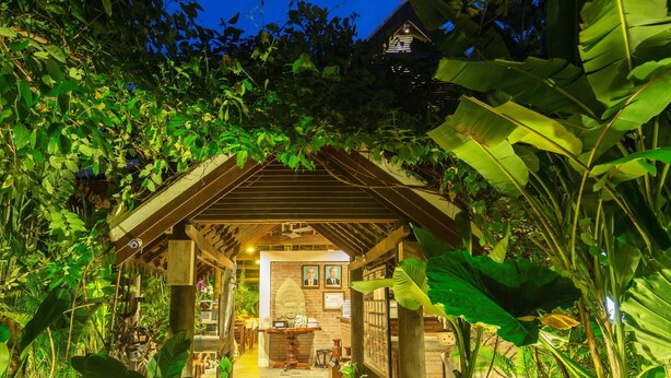 A well-lit pathway leading to a building with a traditional roof, surrounded by lush greenery and potted plants. - Bunwin Residence (Siem Reap)
