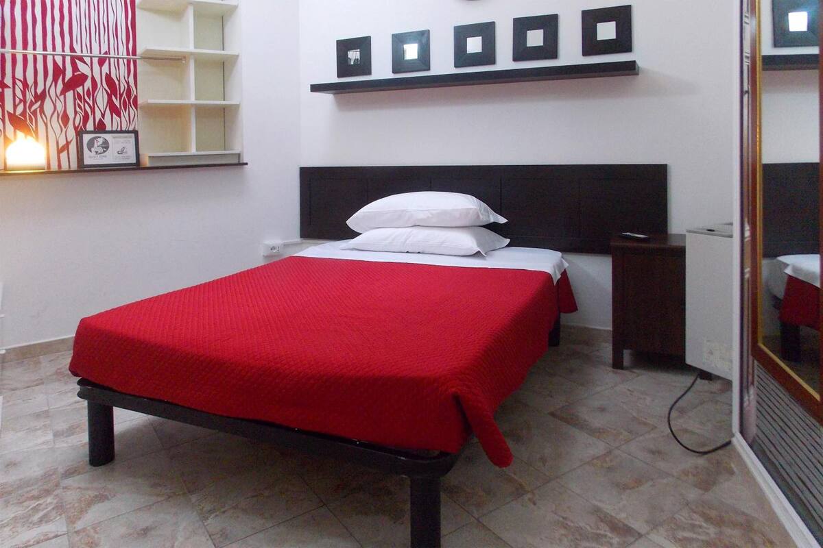 Un dormitorio ordenado con cama, mesita de noche, repisa y estante con fotografías.