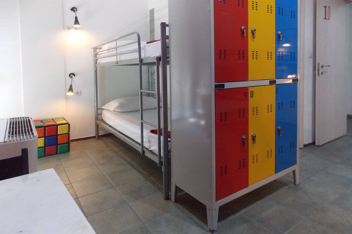 Una cama litera con estructura metálica, un conjunto de taquillas en rojo, amarillo y azul, y un cubo de Rubik en el suelo.