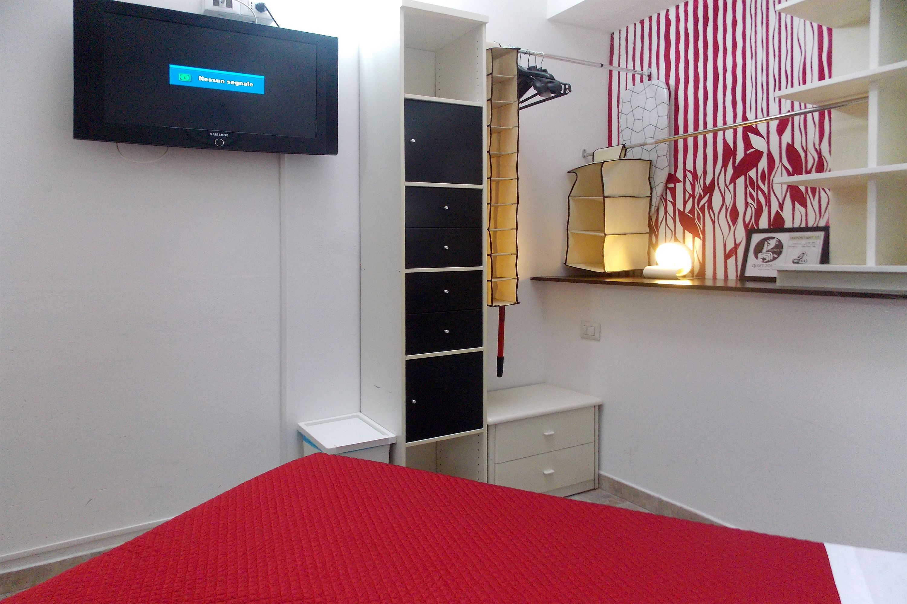 A bedroom with a bed, a TV mounted on the wall, a wardrobe, and a shelf with a lamp.
