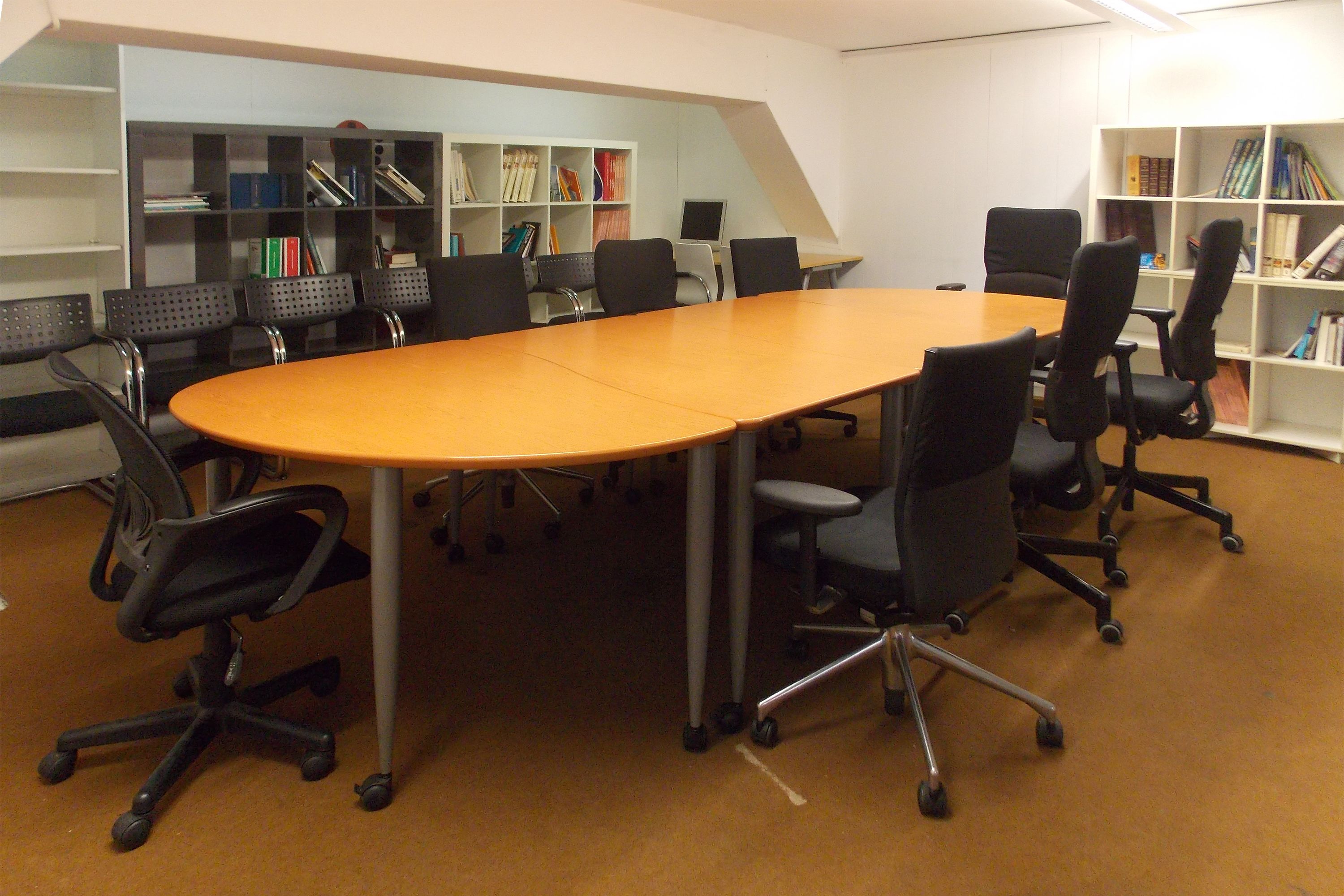 A conference room with a large oval table, six black office chairs, and two white bookshelves filled with books.