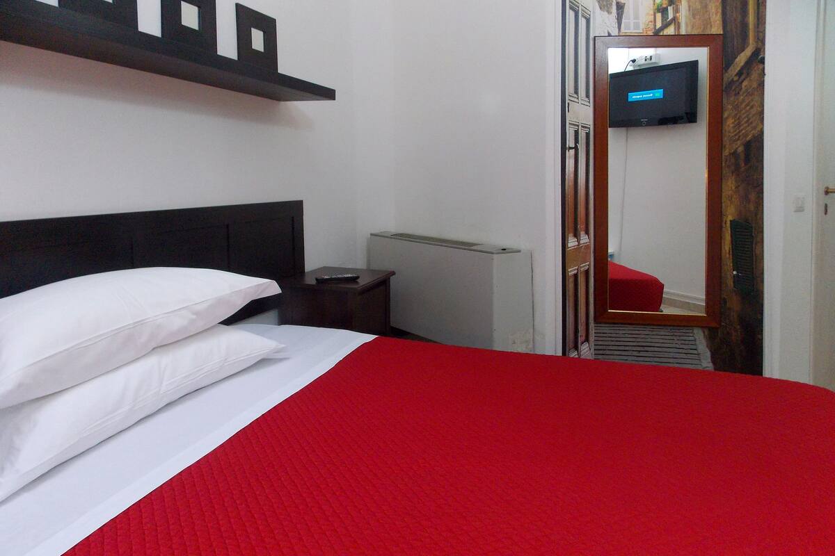 Una habitación de hotel con una cama, una mesita de noche, un calefactor, un televisor y una silla roja.