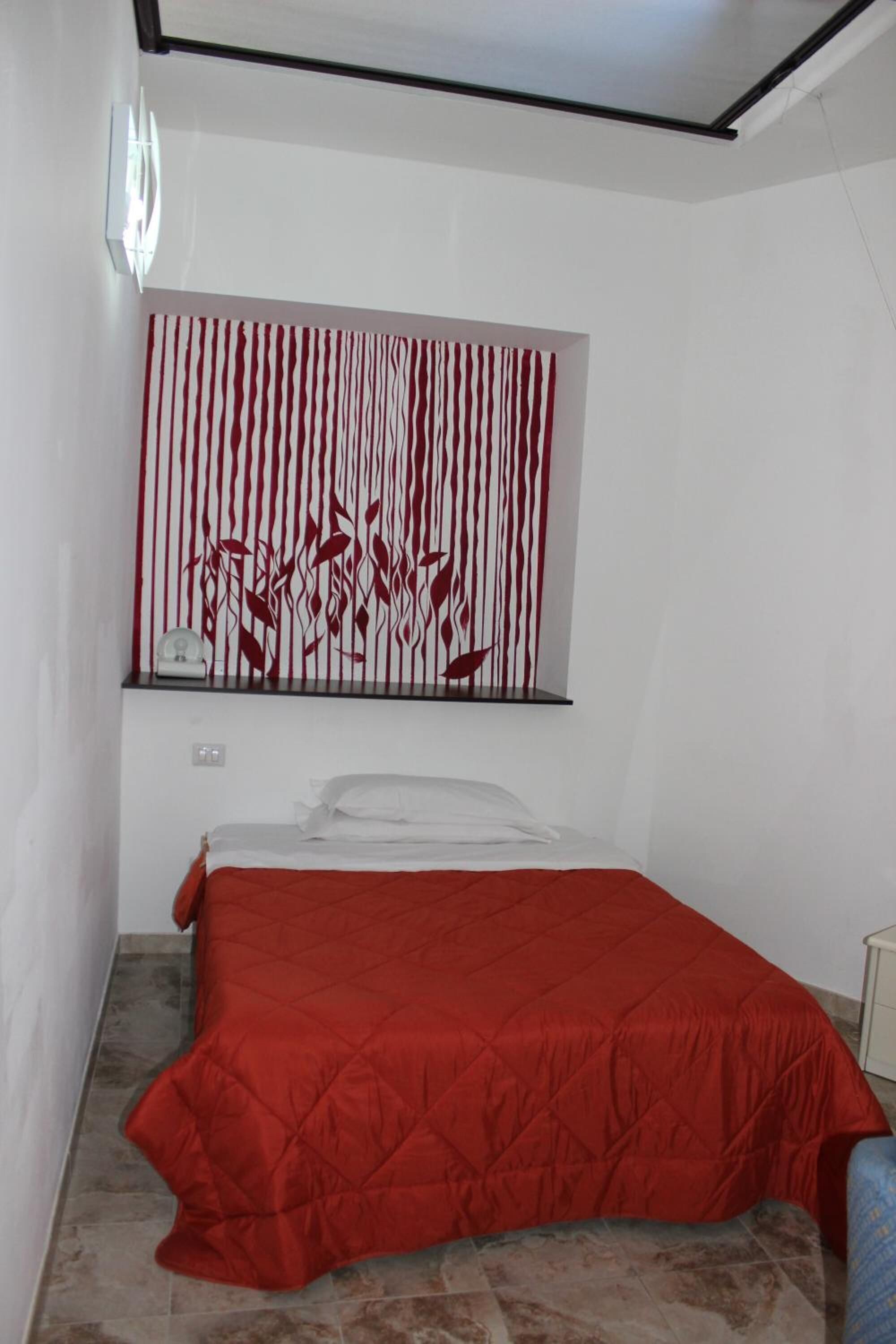 A bedroom with a bed, a red bedspread, a wall-mounted shelf with a clock, and a red and white striped wall decoration.