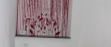 A bedroom with a bed, a red bedspread, a wall-mounted shelf with a clock, and a red and white striped wall decoration.