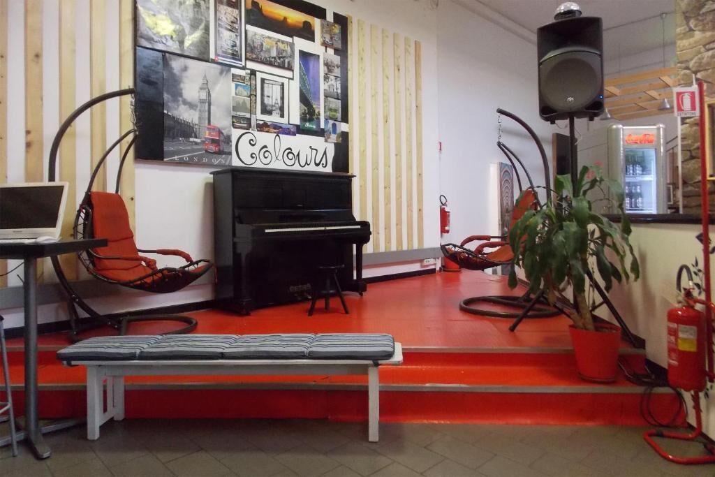 Um quarto com o chão vermelho, um piano preto, um banco, duas cadeiras laranja, um laptop sobre uma mesa e uma parede com quadros emoldurados.