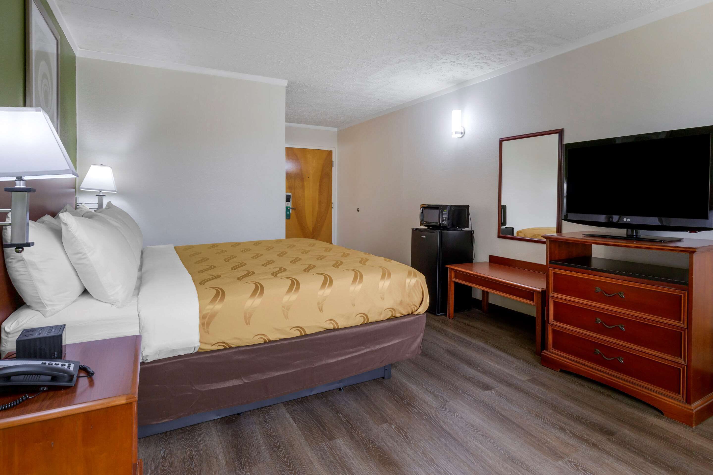 A hotel room with a bed, bedside table with a lamp, a desk with a telephone, a television, and a dresser.