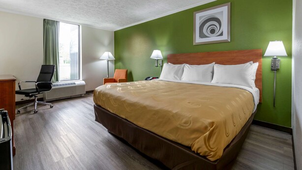 A hotel room with a large bed, two chairs, a desk, and a window with curtains. - Quality Inn - Roxboro South (Roxboro)