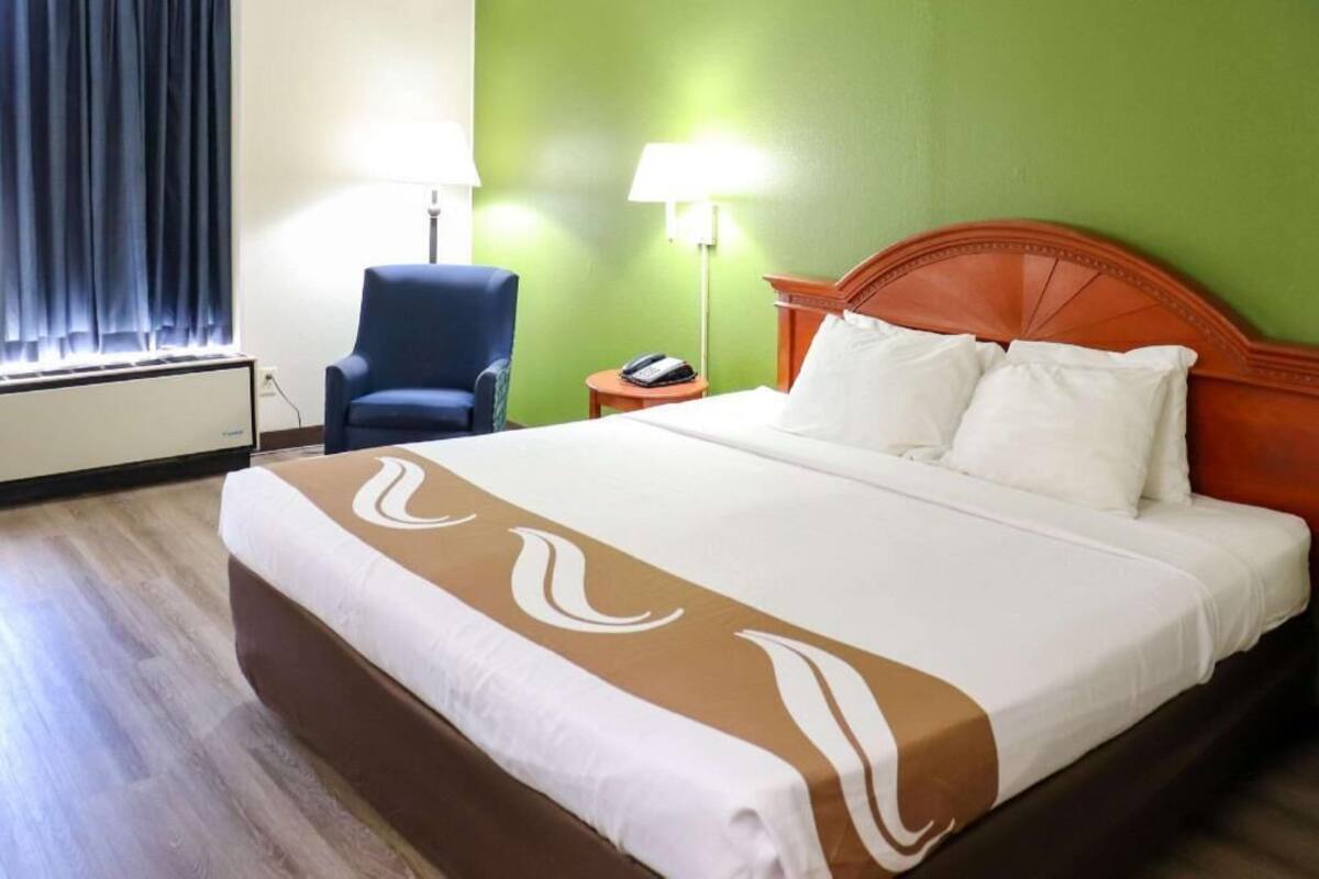 A hotel room with a large bed, a chair, a nightstand with a lamp, and a window with curtains.