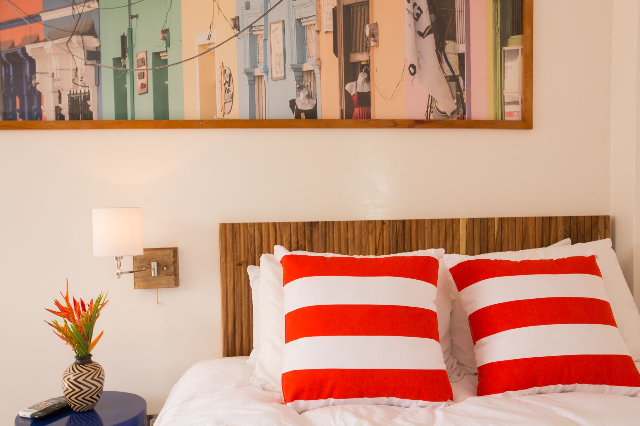 A hotel room with a bed, two red and white striped pillows, a nightstand with a remote control, a vase with flowers, and a wall-mounted lamp.