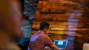 A person working at a wooden desk with a laptop, in a room with wooden walls and pendant lights.