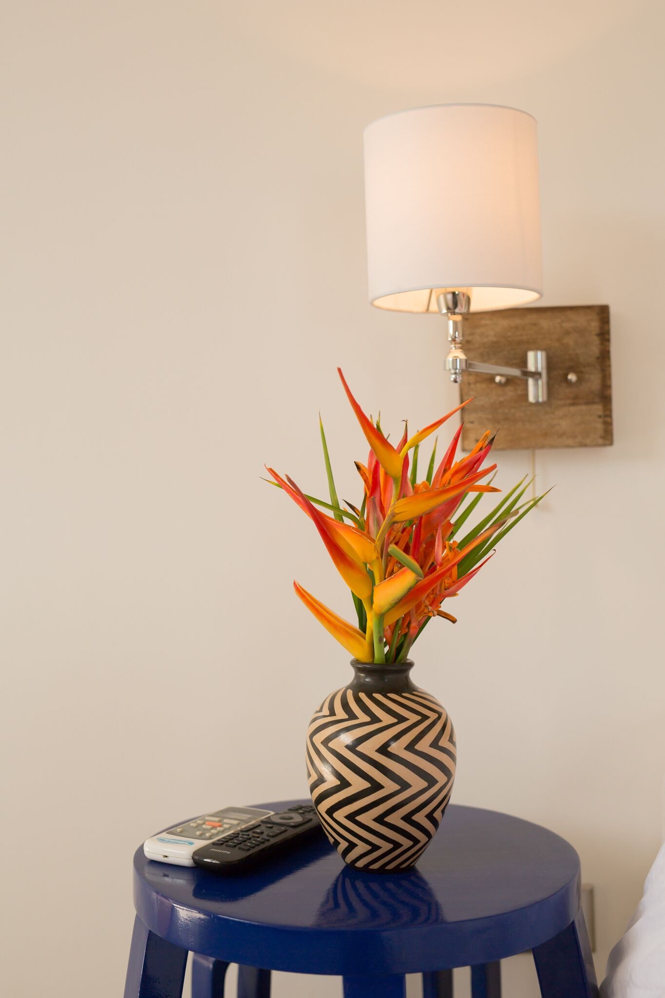 Ein kleiner, blauer Hocker mit einer dekorativen Vase, die eine Pflanze, eine Fernbedienung und eine Wandleuchte enthält.