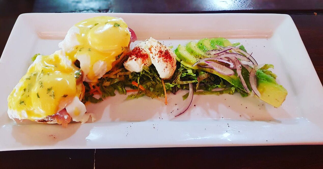En hvid, aflang tallerken med en ret bestående af pocherede æg, hollandaisesauce og en grøn salat med radise og agurk.
