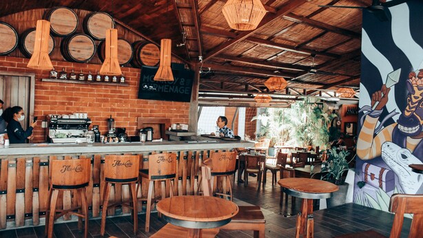A rustic bar area with wooden chairs and tables, a brick wall with hanging light fixtures, and a mural on the wall.
