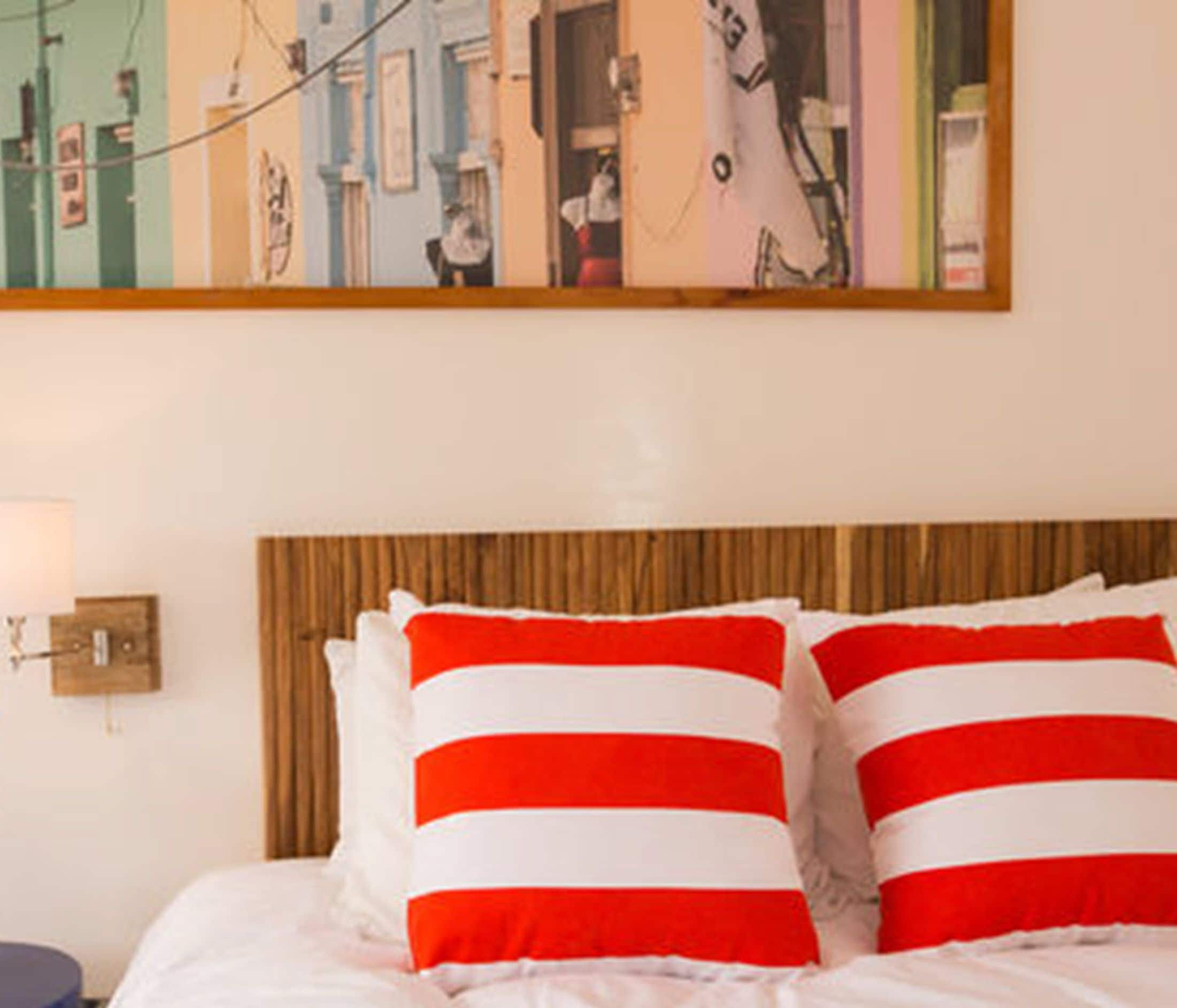 A hotel room with a bed featuring red and white striped pillows, a wooden headboard, and a wall-mounted lamp.