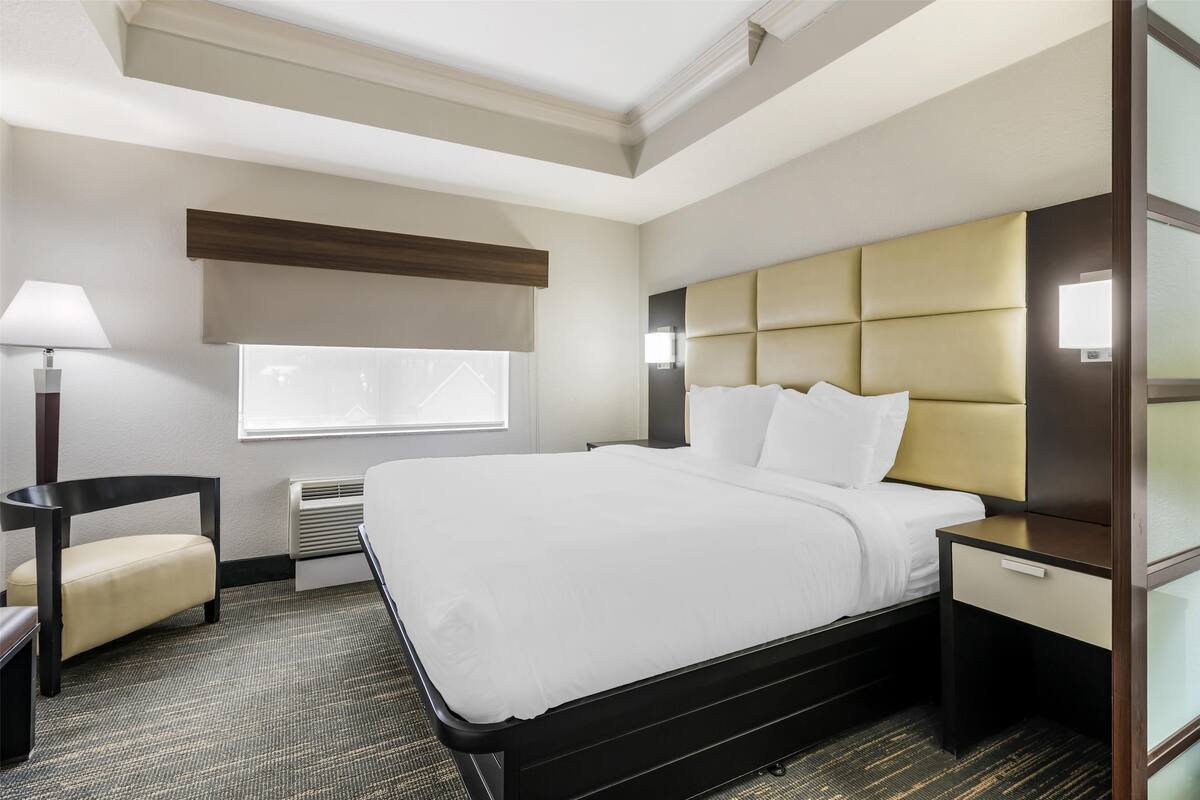 A hotel room with a large bed, a bedside table, a lamp, a chair, and a window with blinds.