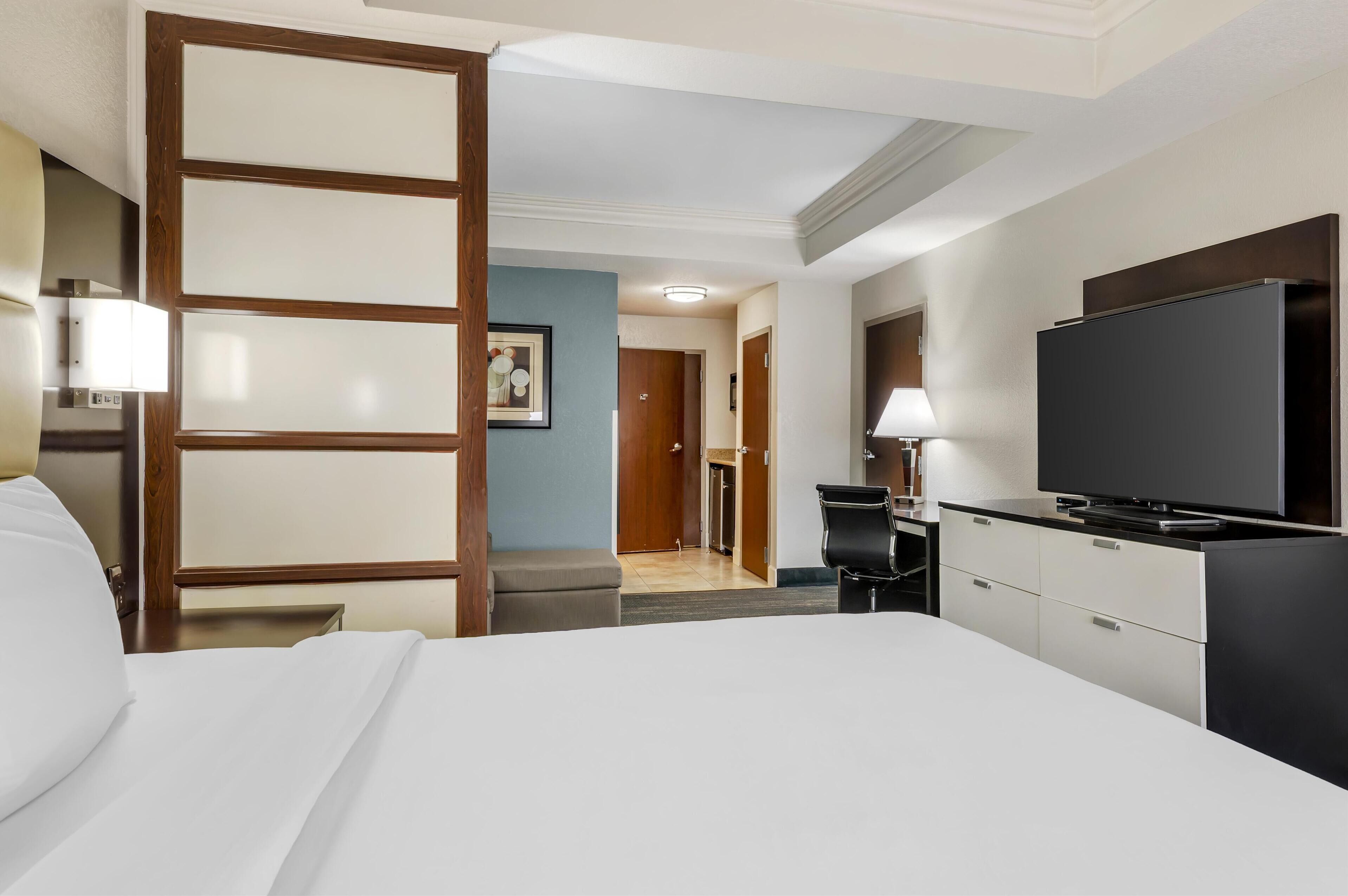 Quarto de hotel com cama, escrivaninha, televisão e porta que dá para um corredor.