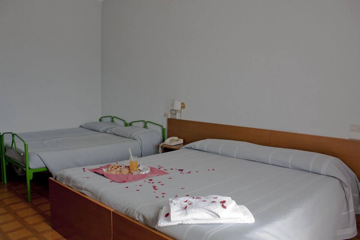A hotel room with two single beds, a wooden headboard, a bedside table with a phone, and a small table with a drink and rose petals.