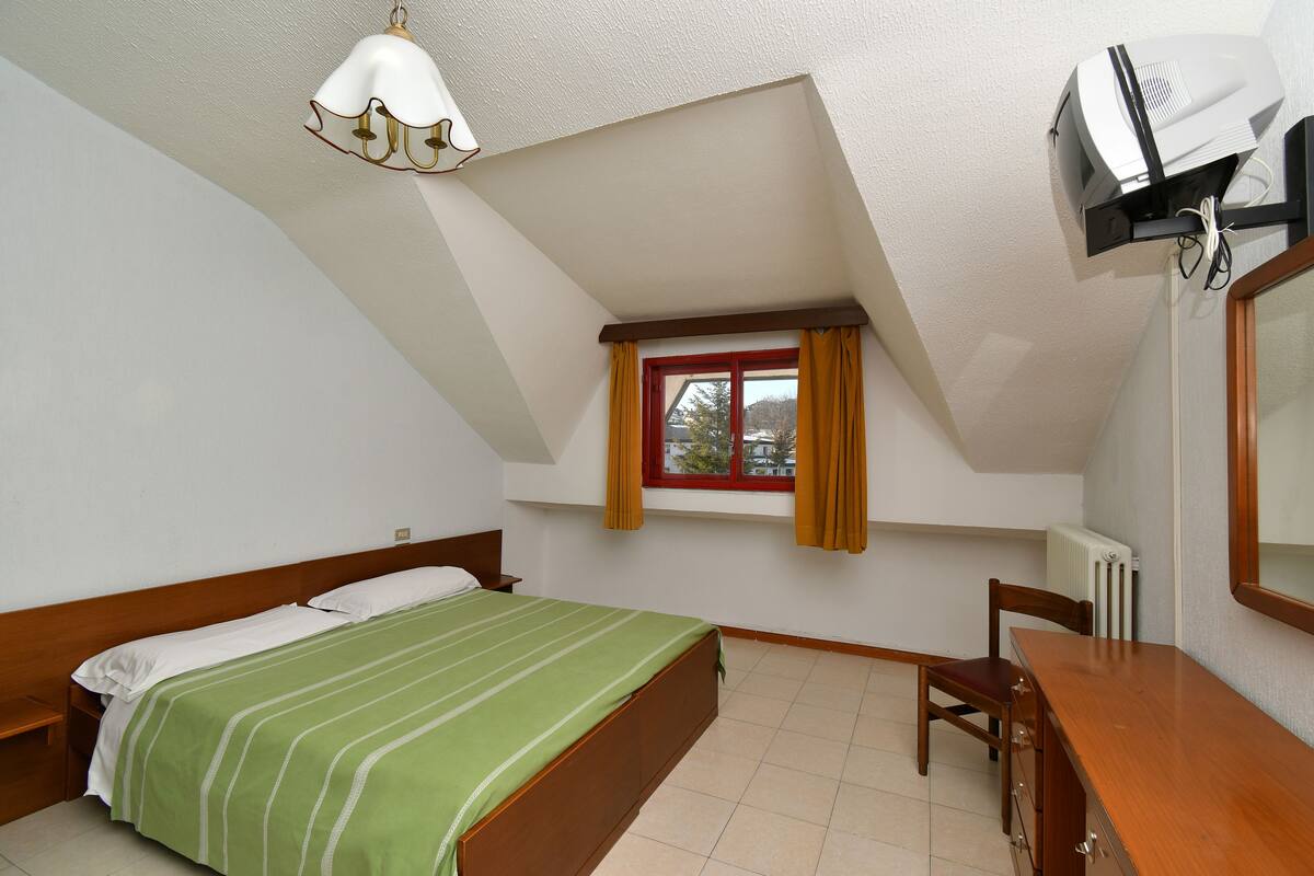 A bedroom with a bed, a desk, a chair, a window with curtains, a radiator, and a ceiling fan.