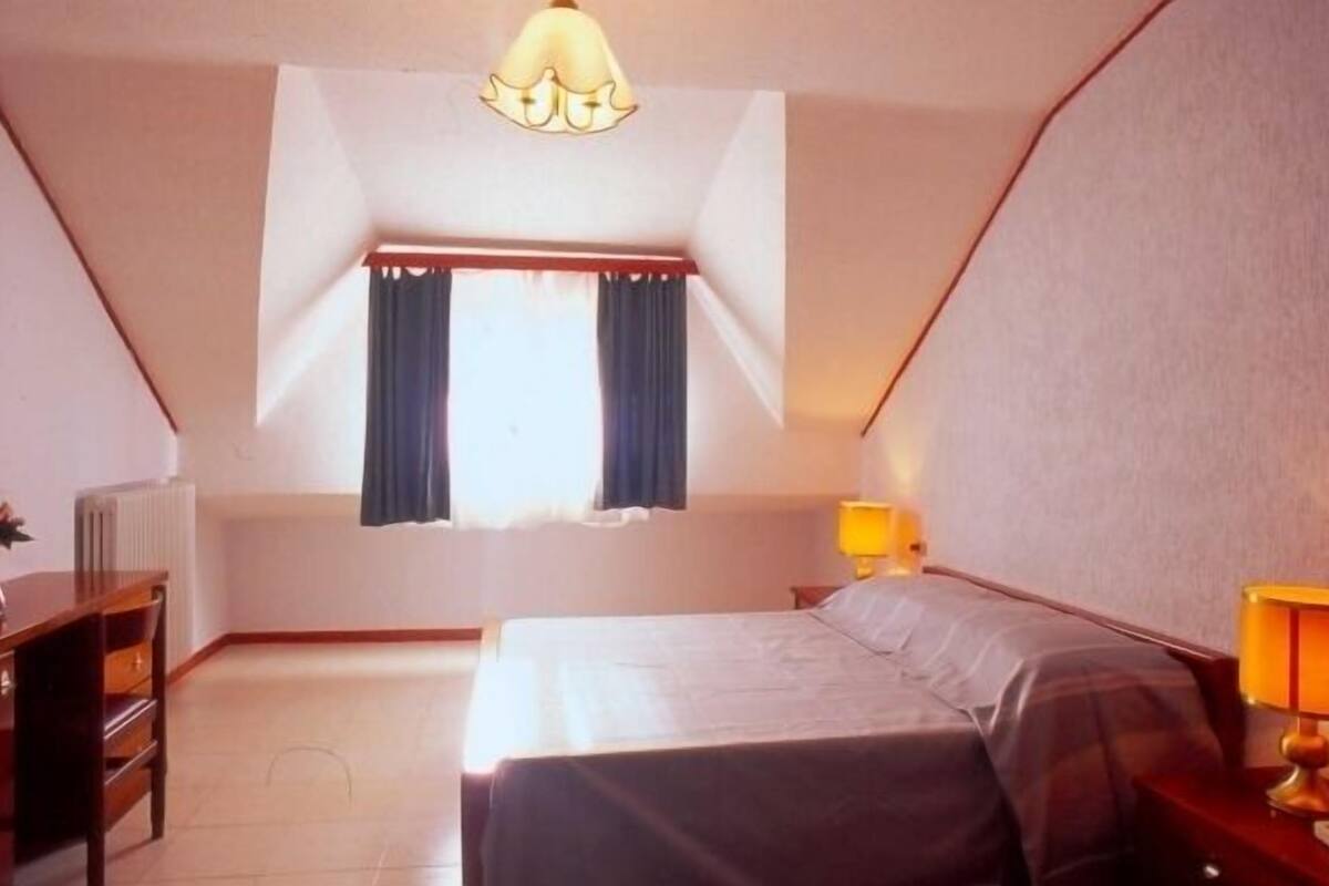 A bedroom with a bed, a desk, a chair, a lamp, and a window with curtains.