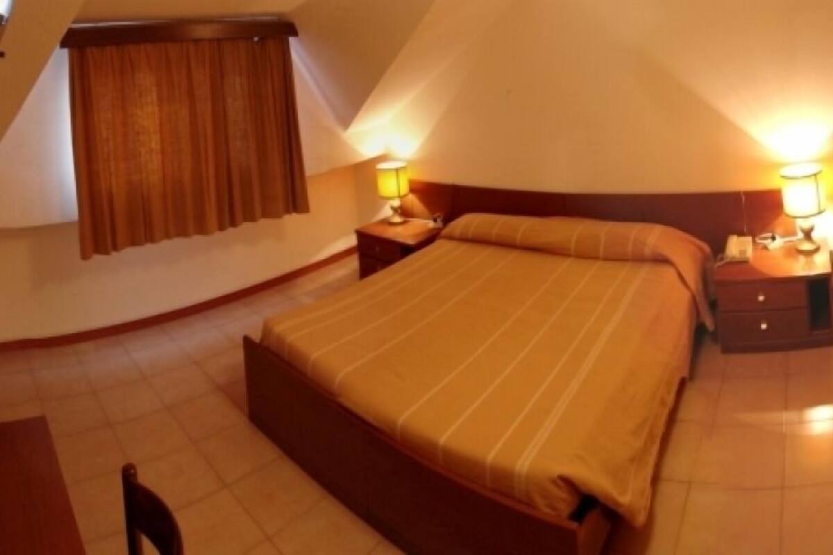 A bedroom with a large bed, two bedside tables with lamps, a window with curtains, and a small table with a vase of flowers.
